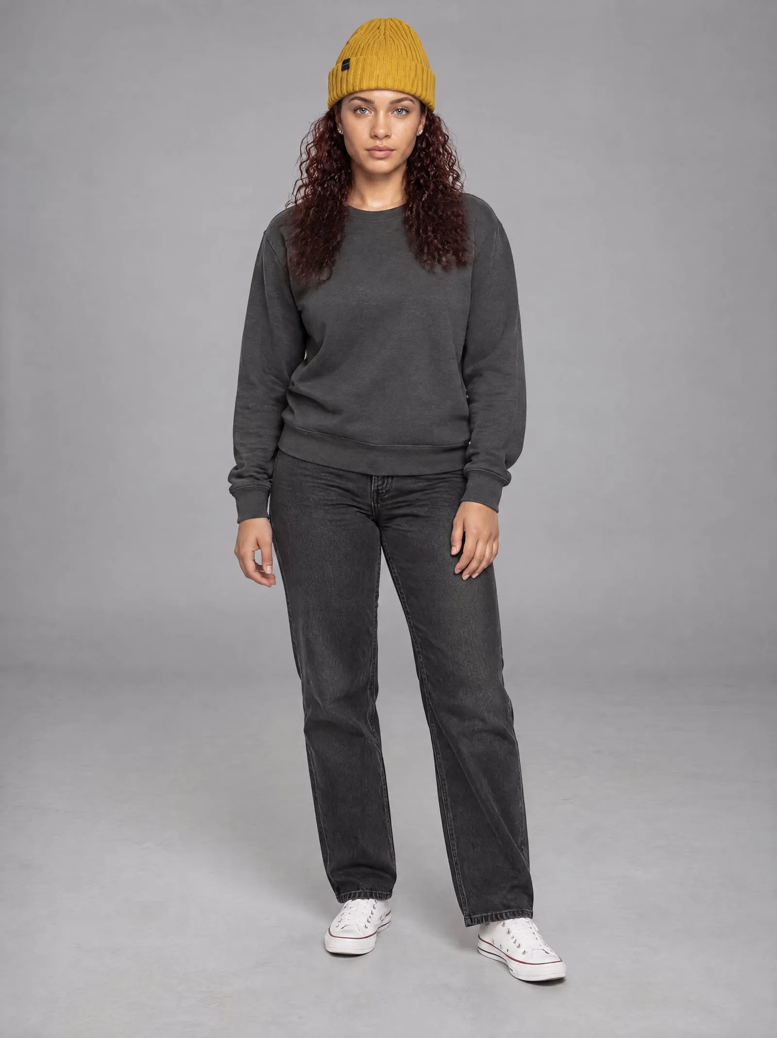 Noor wearing the mustard ribbed beanie, standing, in a grey studio