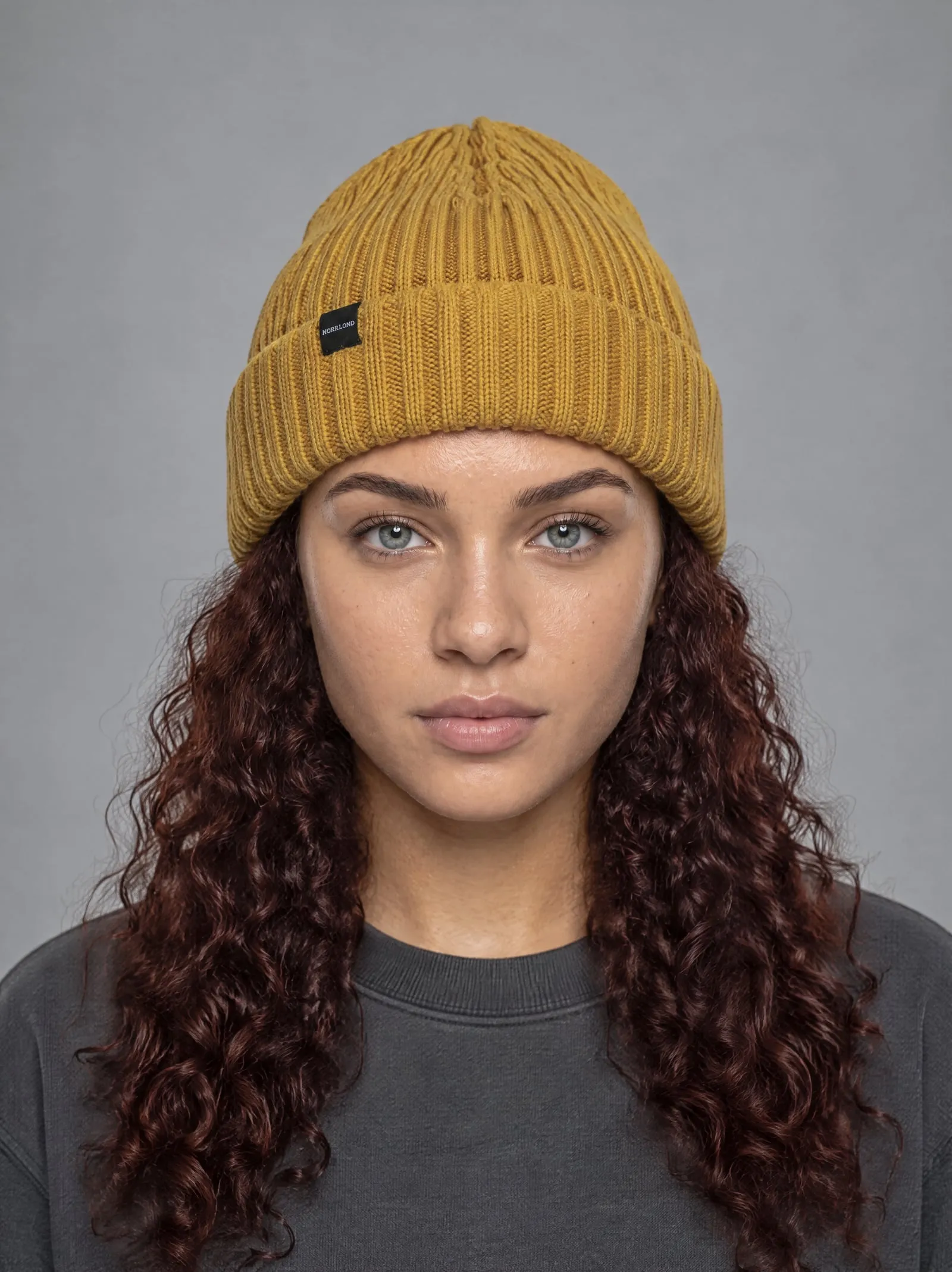 Noor wearing the mustard ribbed beanie, in a portrait, in a grey studio