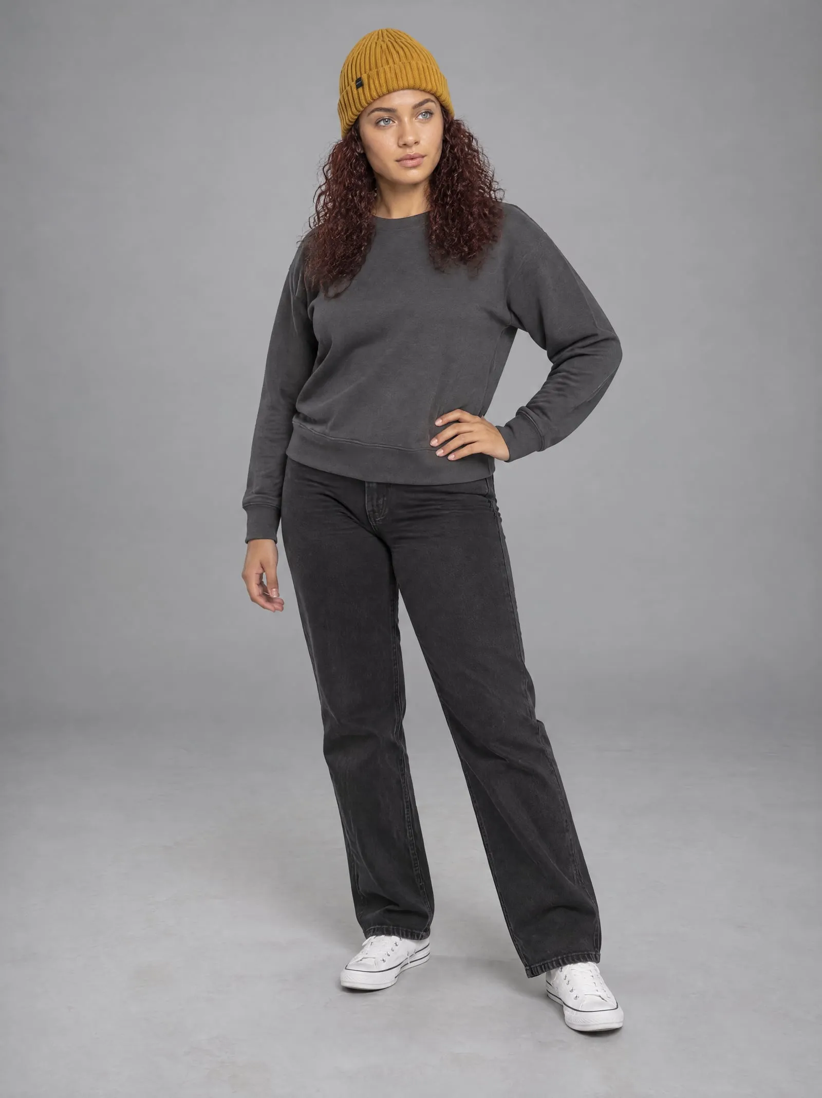 Noor wearing the mustard ribbed beanie, hand on hip, in a grey studio