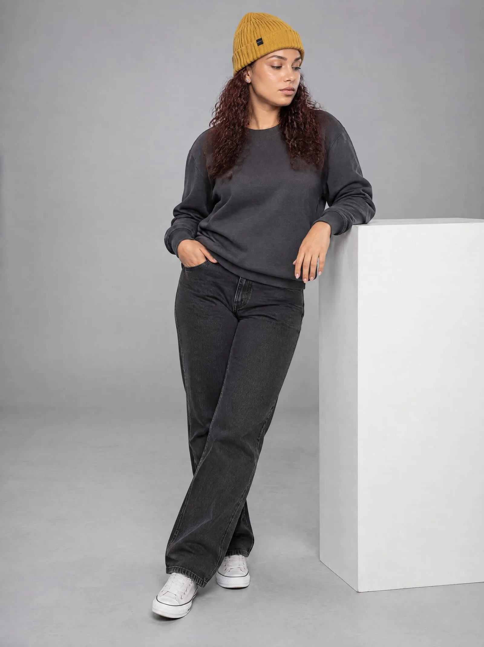 Noor wearing the mustard ribbed beanie, leaning, in a grey studio