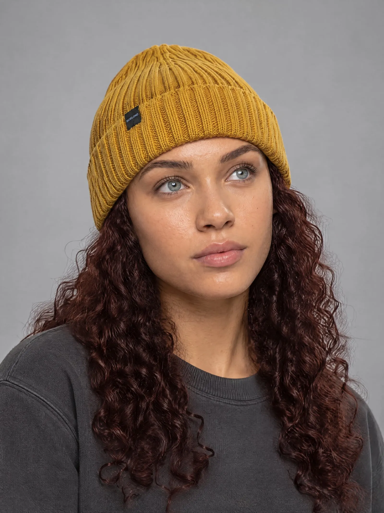 Noor wearing the mustard ribbed beanie, chin lifted, in a grey studio
