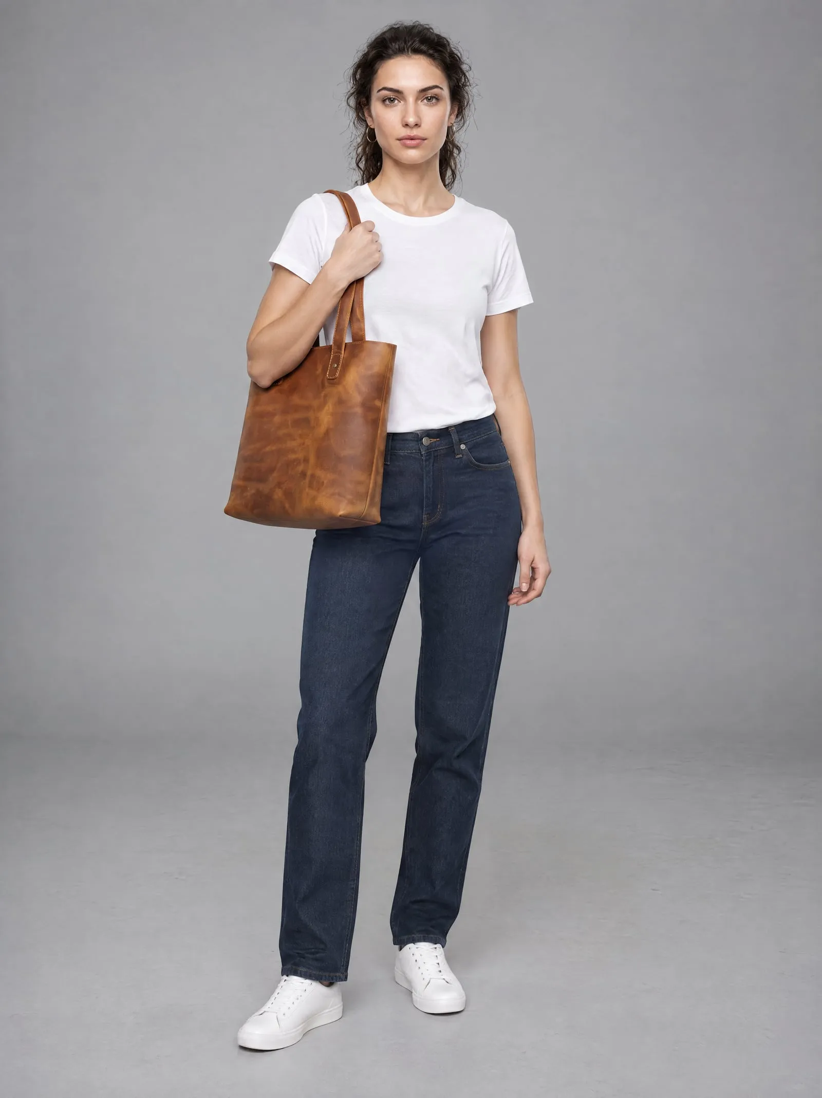 Mira wearing the tan leather tote bag, standing, in a grey studio