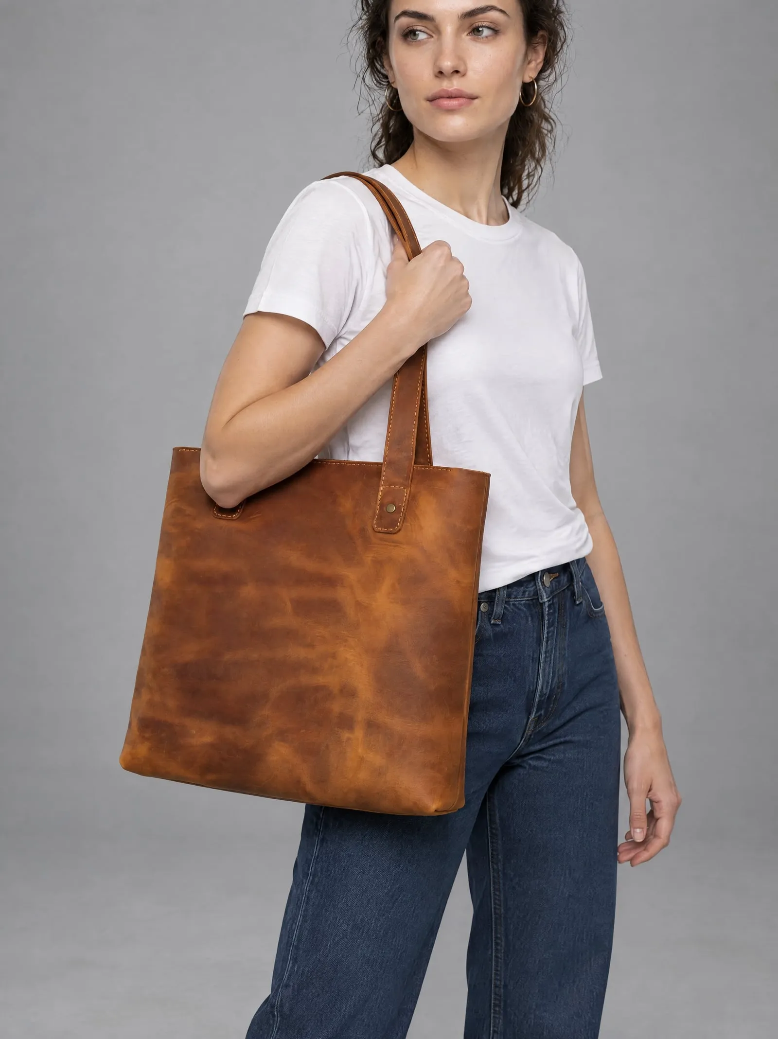 Mira wearing the tan leather tote bag, holding the strap, in a grey studio