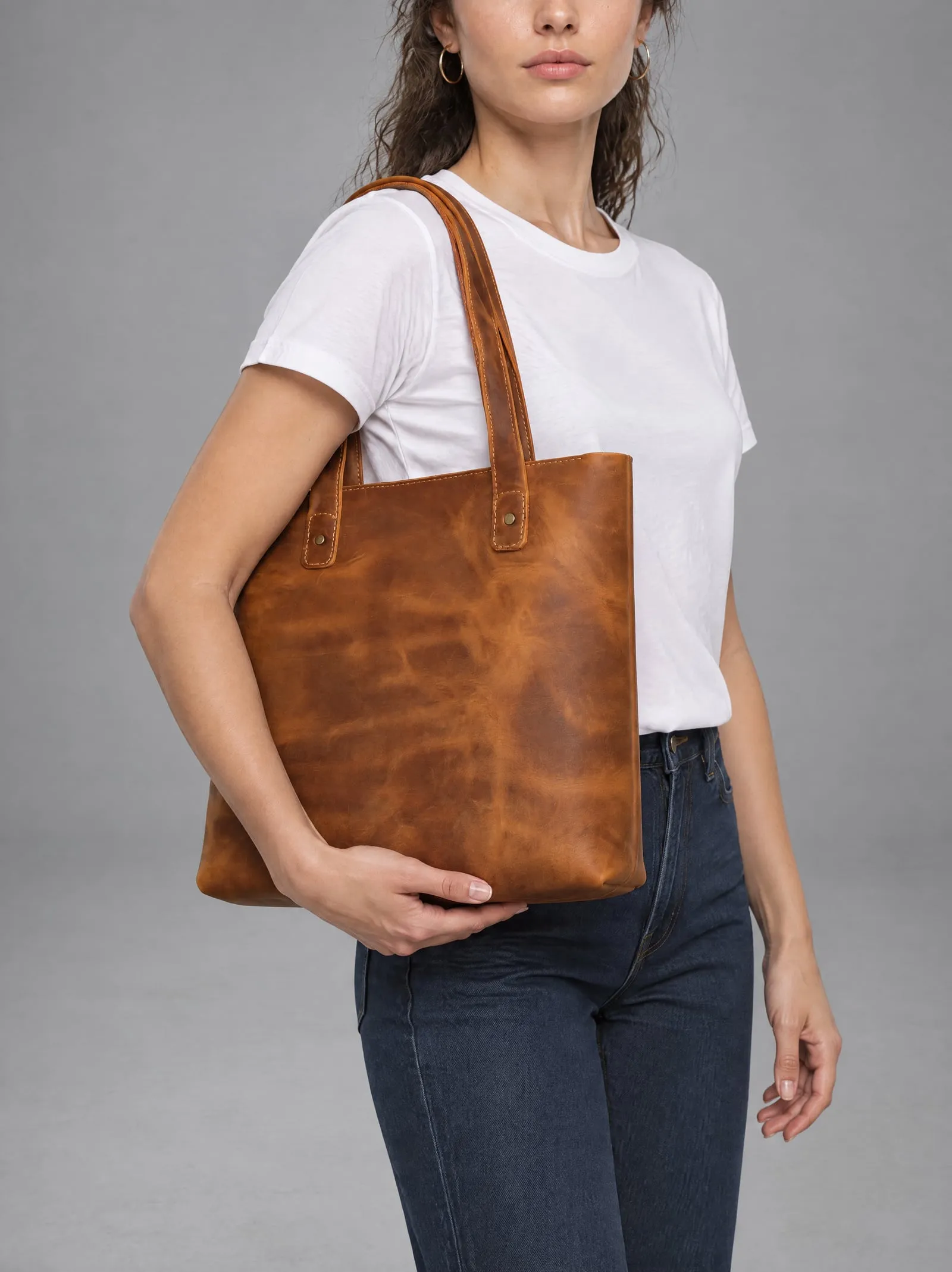 Mira wearing the tan leather tote bag, holding the bag in front of her, in a grey studio