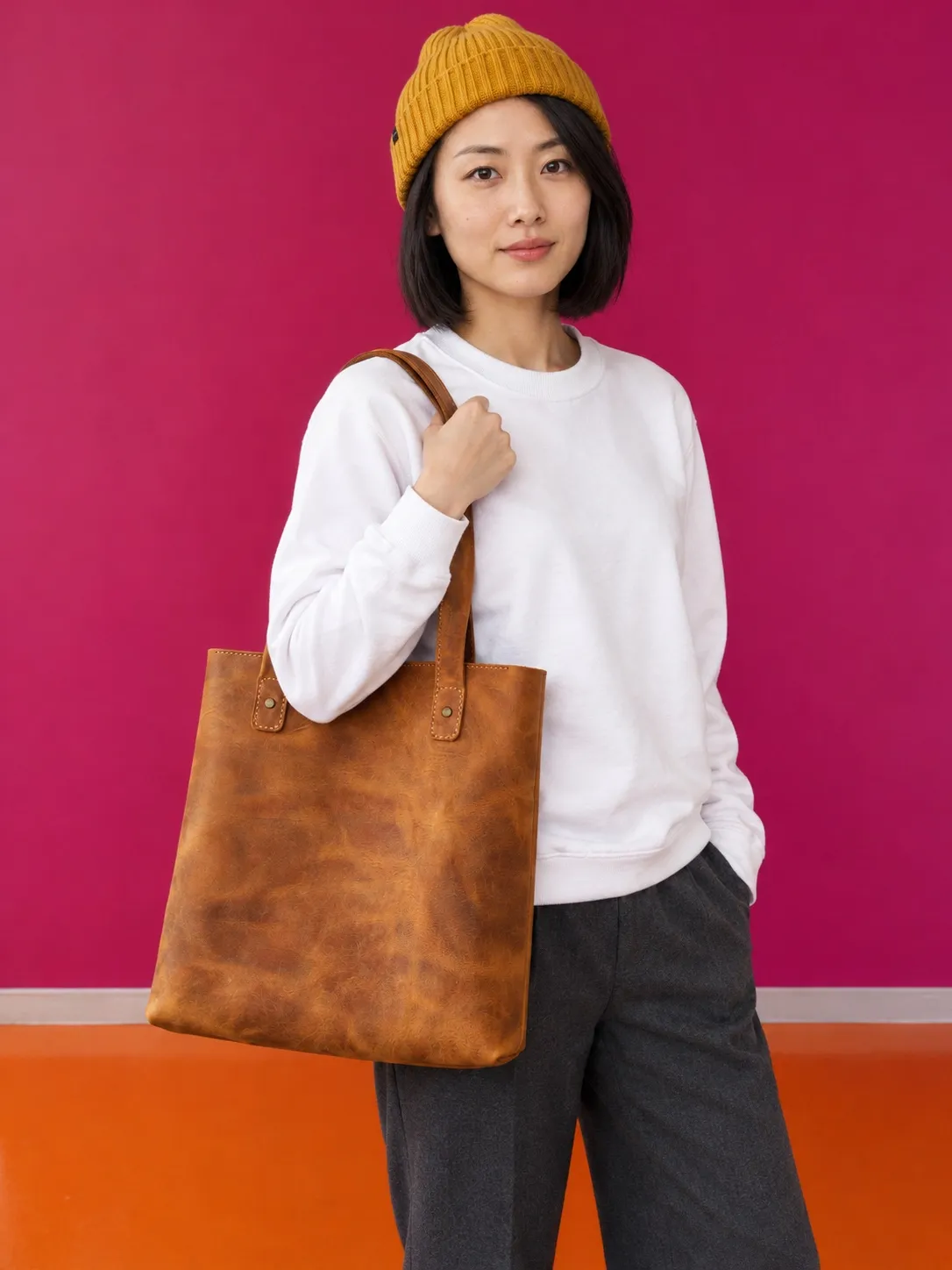 Hana in a white sweater and a mustard beanie, a tan leather tote on her shoulder, against a magenta wall